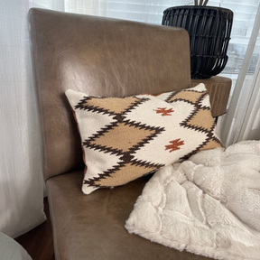 A Handwoven Blazheny (Blissful) Pillow and a white blanket rest on a brown leather chair near a window with sheer curtains.