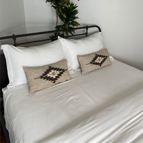 A neatly made bed with white linens, two pillows, and two decorative cushions, including the stylish Handwoven Chitki (Crisp) Pillow.