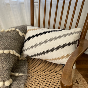 A Handwoven Promin (Beam) Wool Pillow and chunky knit blanket rest on a wooden rocking chair with a woven seat.
