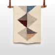 The Harmonia A Wool Statement Throw / Baby Blanket features woven geometric triangles in blue, grey, brown, and beige on a wooden rod for a cozy, stylish accent.