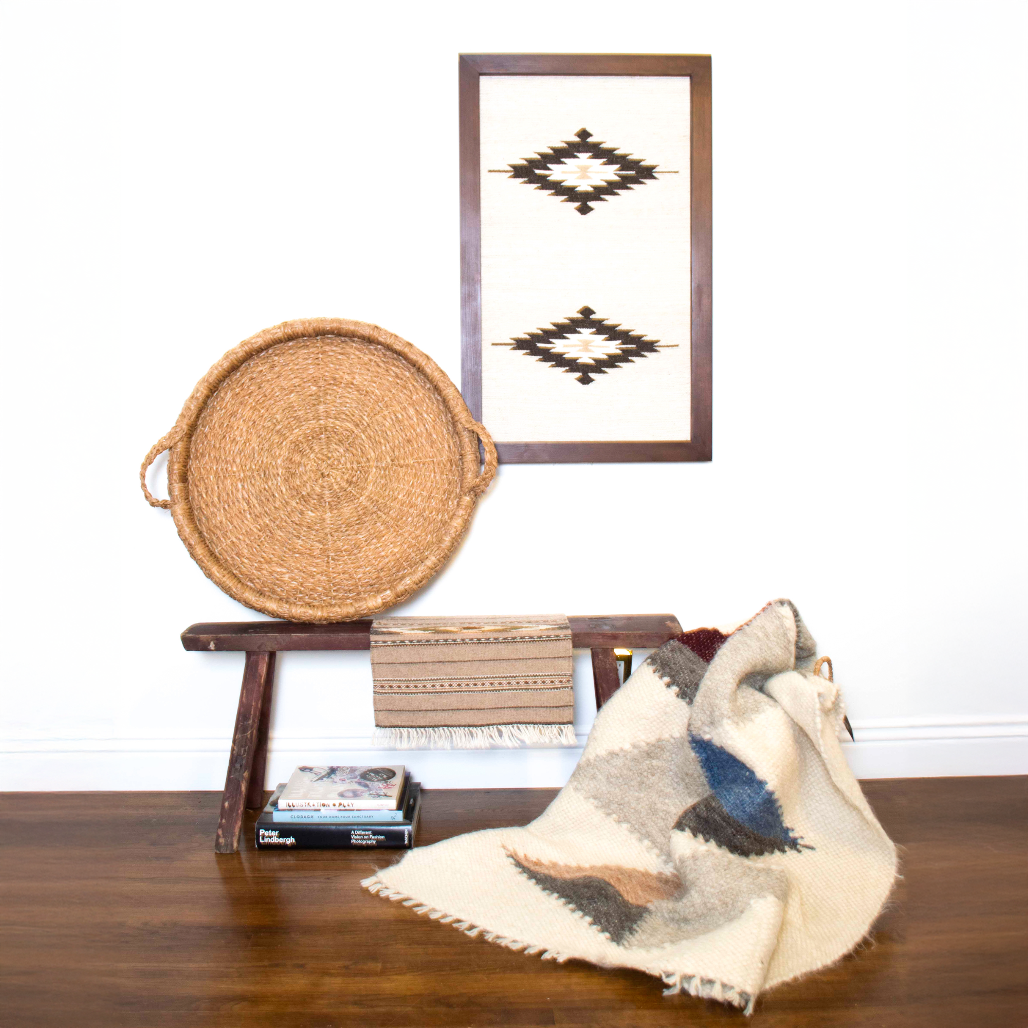 The Harmonia A (Harmony) Wool Statement Throw drapes over a wooden bench, paired with a woven tray and patterned textile beneath framed geometric wall art for a cozy, modern look.