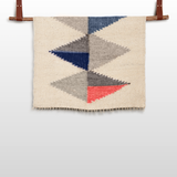 The Harmonia B (Harmony) Wool Statement Throw features blue, gray, and red geometric triangles on a beige background, inspired by modern art, and doubles as an elegant wall hanging displayed on a wooden rod.