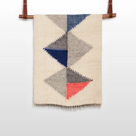 The Harmonia B (Harmony) Wool Statement Throw / Baby Blanket features colored triangles in blue, gray, and coral on a cream background, inspired by modern art blanket designs and displayed on a wooden rod.