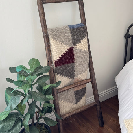 The Harmonia A (Harmony) Wool Statement Throw / Baby Blanket with a geometric-knit design is draped over a wooden ladder beside a green potted plant and a bed.