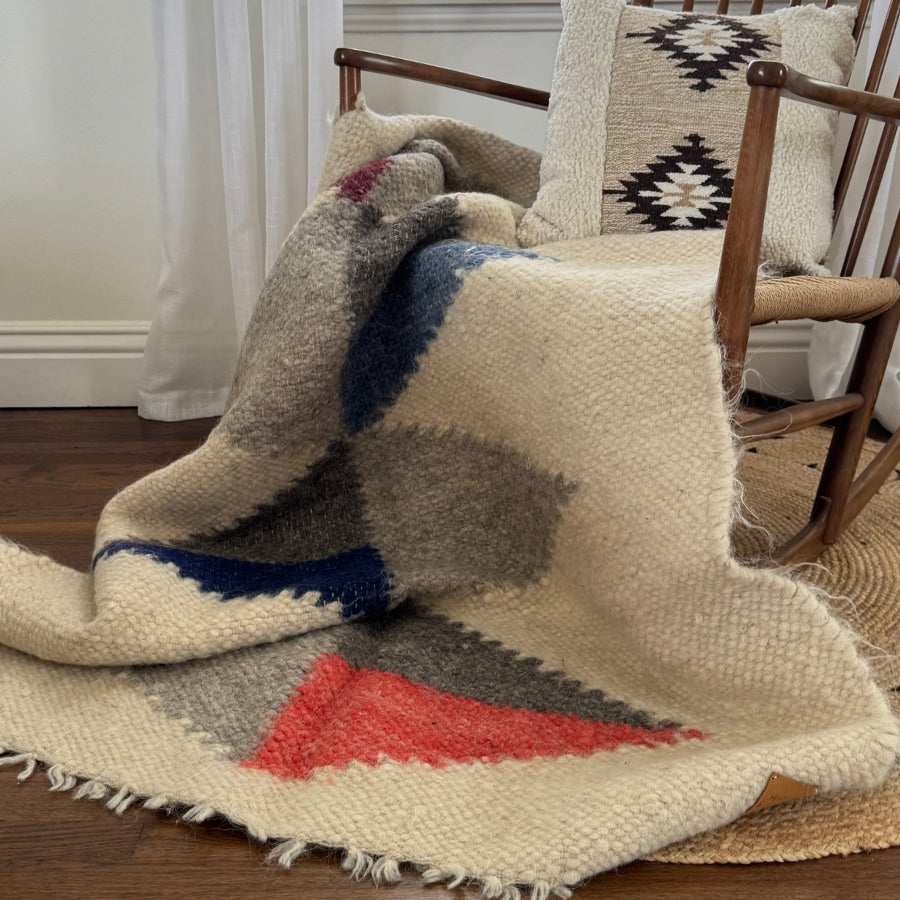 The Harmonia B (Harmony) Wool Statement Throw / Baby Blanket, in cream with red, blue, and gray geometric patterns, is draped over a wooden chair with a patterned cushion.