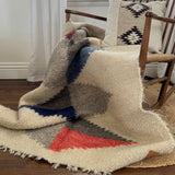 The Harmonia B (Harmony) Wool Statement Throw / Baby Blanket, in cream with red, blue, and gray geometric patterns, is draped over a wooden chair with a patterned cushion.
