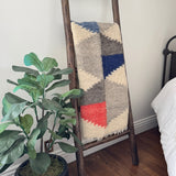 The Harmonia B (Harmony) Wool Statement Throw/Baby Blanket with a modern art design drapes over a wooden ladder beside a potted plant and a bed with white bedding.