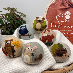 Six Fluff Ewes "Hedgehog & Homies" 100% premium wool dryer balls with woodland designs rest on a white cloth by a plant and bag—an eco-friendly, natural fabric softener for your laundry.