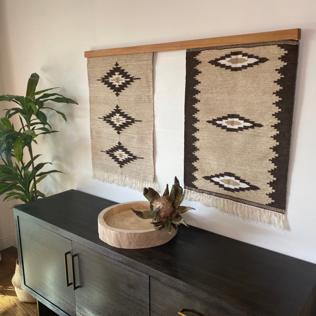 Two Heredij (Heritage) wool wall hangings with geometric patterns, reminiscent of Carpathian mountain rugs, displayed above a dark cabinet adorned with a plant and wooden decor.