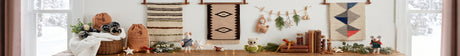 A festive tabletop with holiday decor, woven wall hangings, gift bags, books, and small animal figurines.