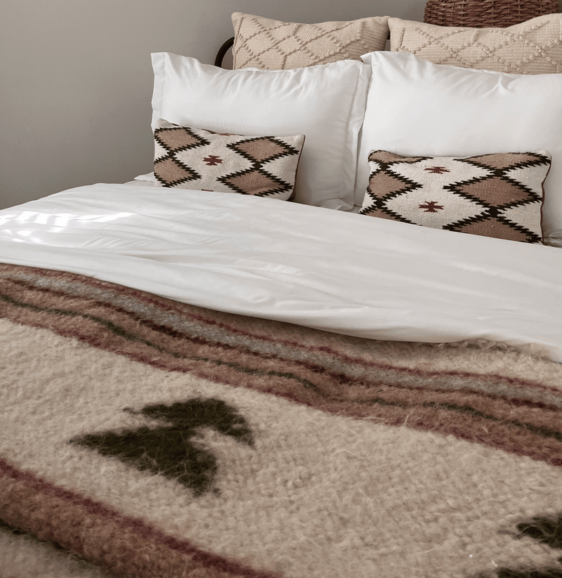 Cozy bed with white sheets, two patterned pillows, and a textured wool blanket with earthy tones.