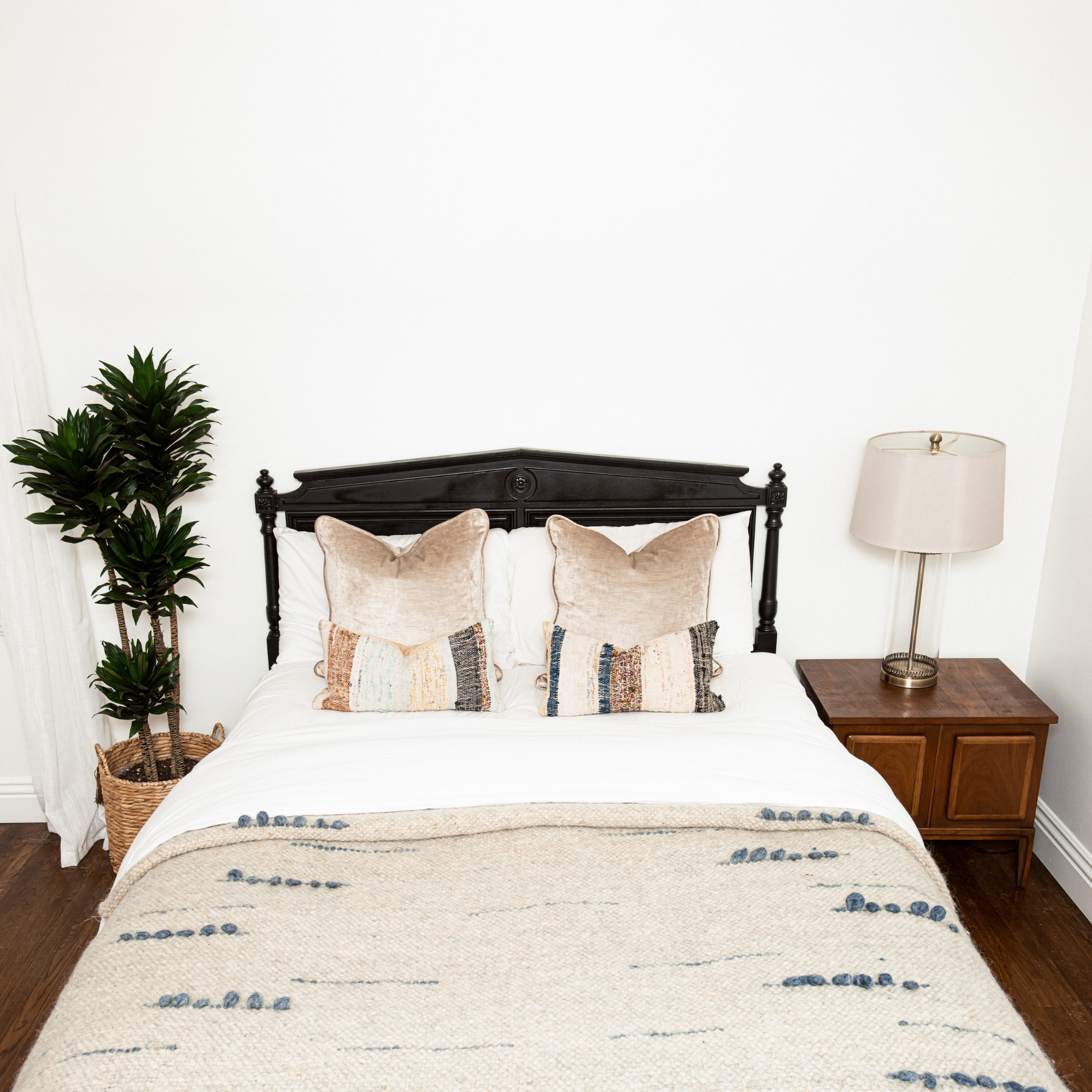 A neatly made bed with two pillows, the Horoshy (Kind) Wool Statement Bed Blanket crafted by Ukrainian weavers, a potted plant, and a lamp on a wooden nightstand.