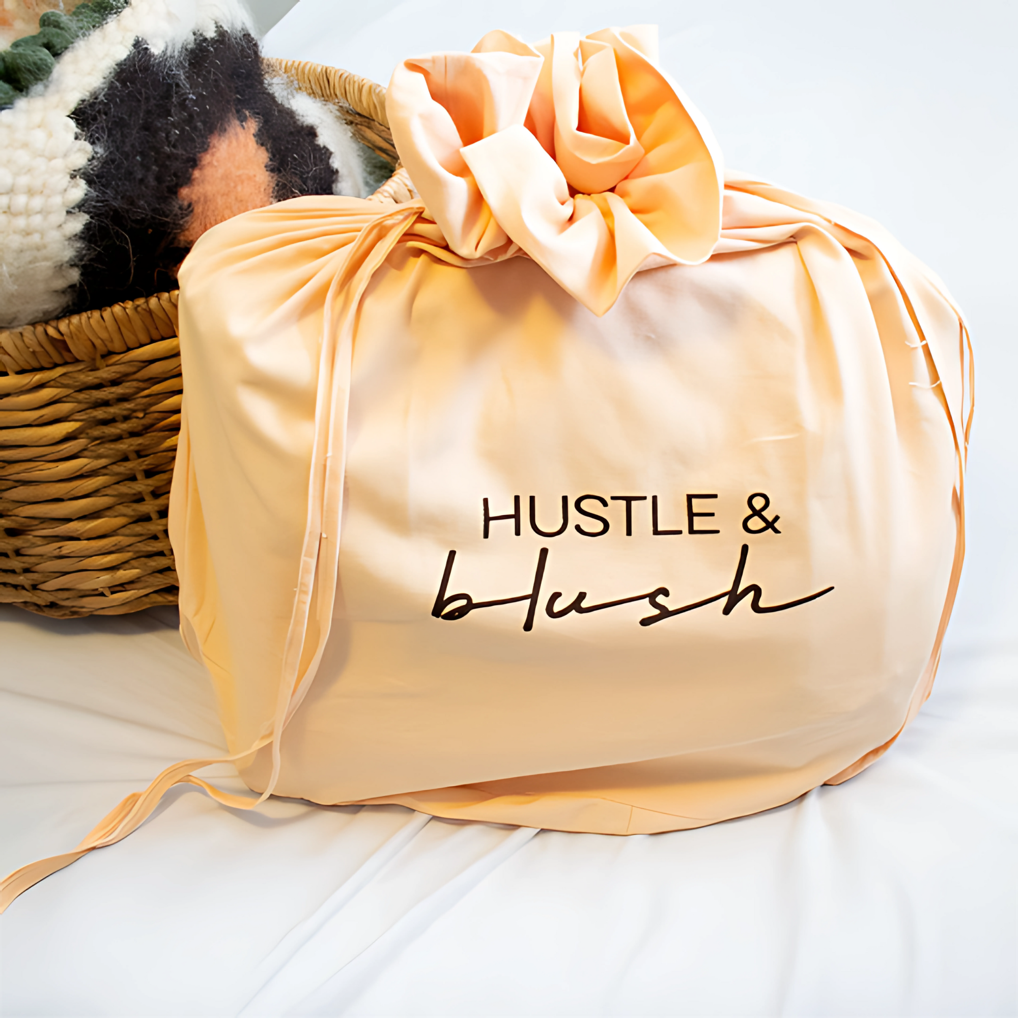 Peach fabric bag with "HUSTLE & blush" text sits next to a woven basket and the Horoshy (Kind) Wool Statement Bed Blanket, inspired by Ukrainian weavers, all on a white surface.