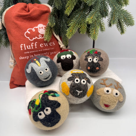 The Fluff Ewes Premium XL Dryer Balls "Sheep in Heavenly Peace" Holiday Limited Edition 6 pack features eco-friendly wool balls with cute animal faces, displayed under pine branches with a festive red “fluff ewes” bag.