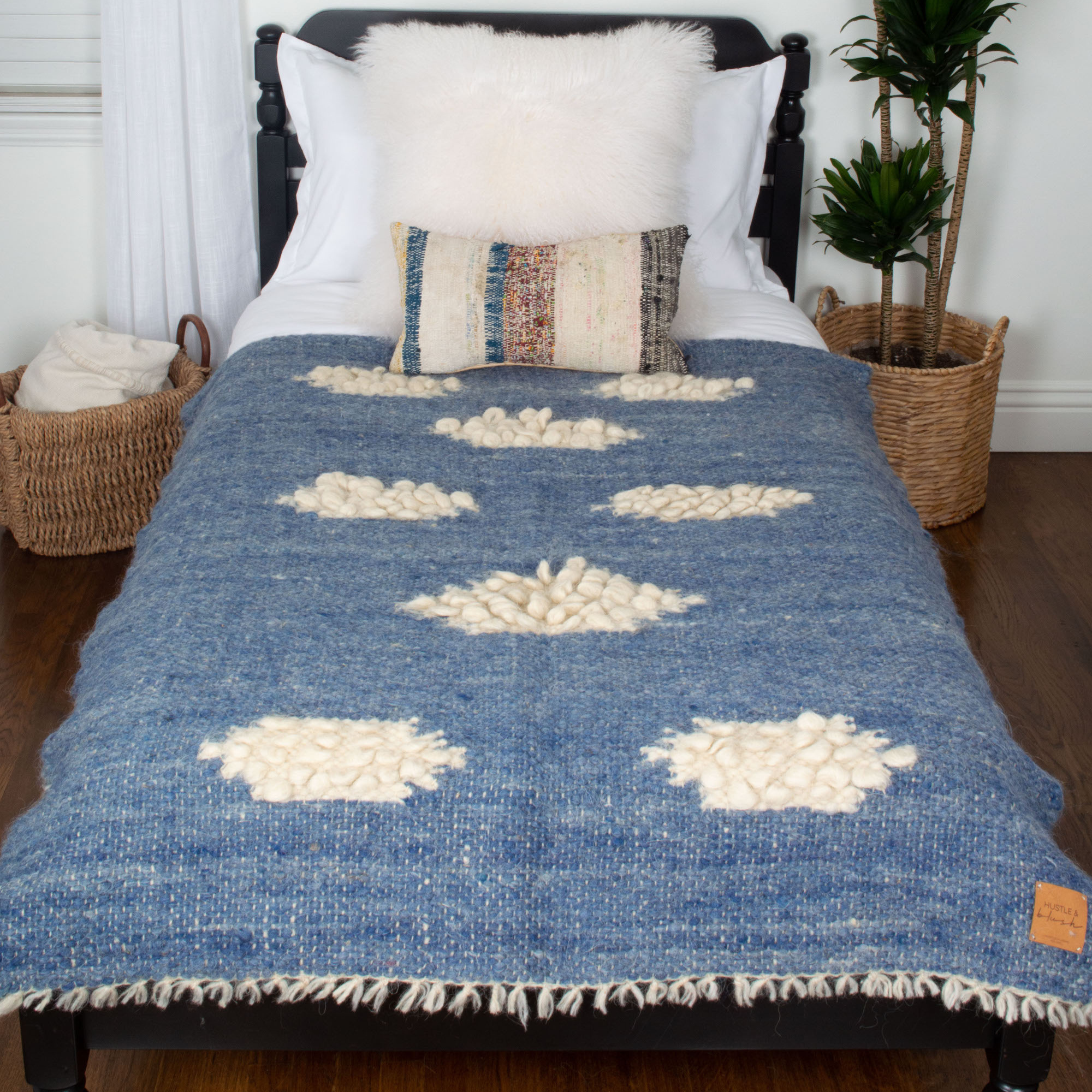 A bed with the Khmara (Cloud) Wool Statement Throw featuring white cloud patterns, a fluffy pillow, and baskets beside it on a wooden floor.