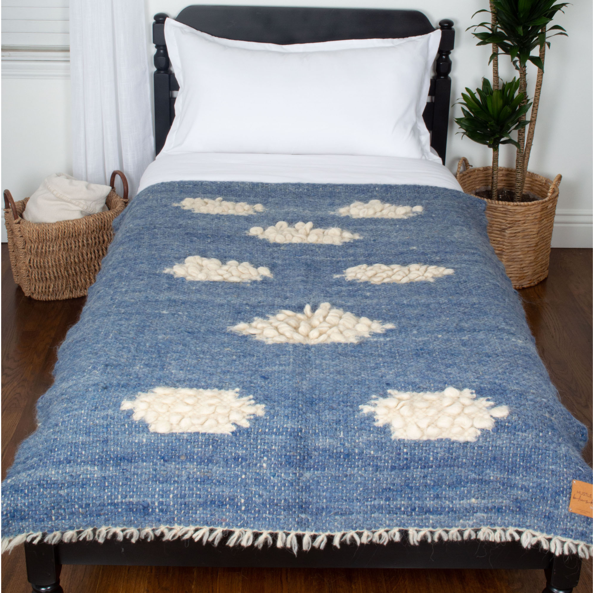 The Khmara (Cloud) Wool Statement Throw / Baby Blanket, in blue and white tufted wool, adds texture to a twin bed, complemented by nearby woven baskets and plants.