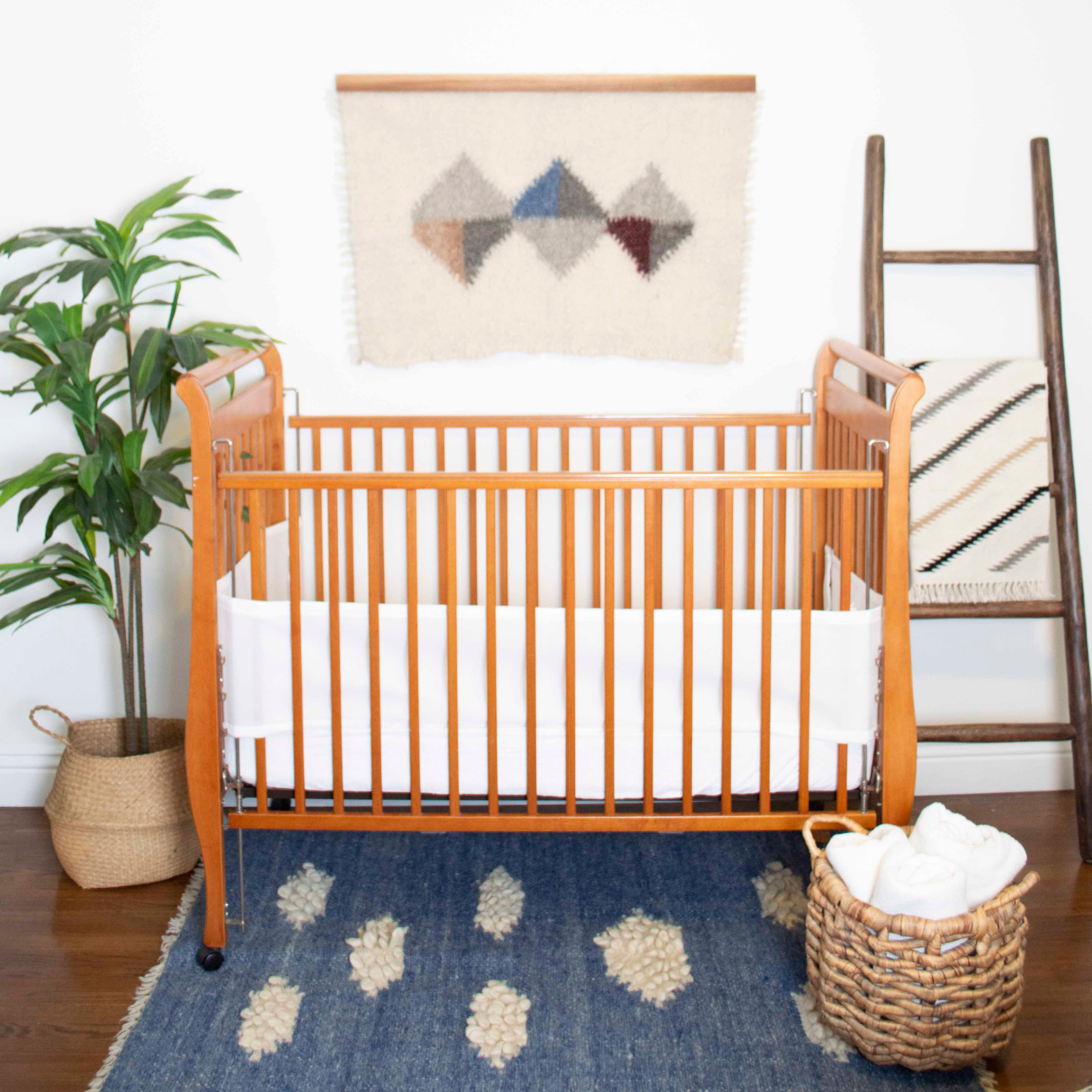A wooden crib with white bedding and the Khmara (Cloud) Wool Statement Throw / Baby Blanket on a blue rug, beside a plant, ladder, and basket in a cozy nursery.