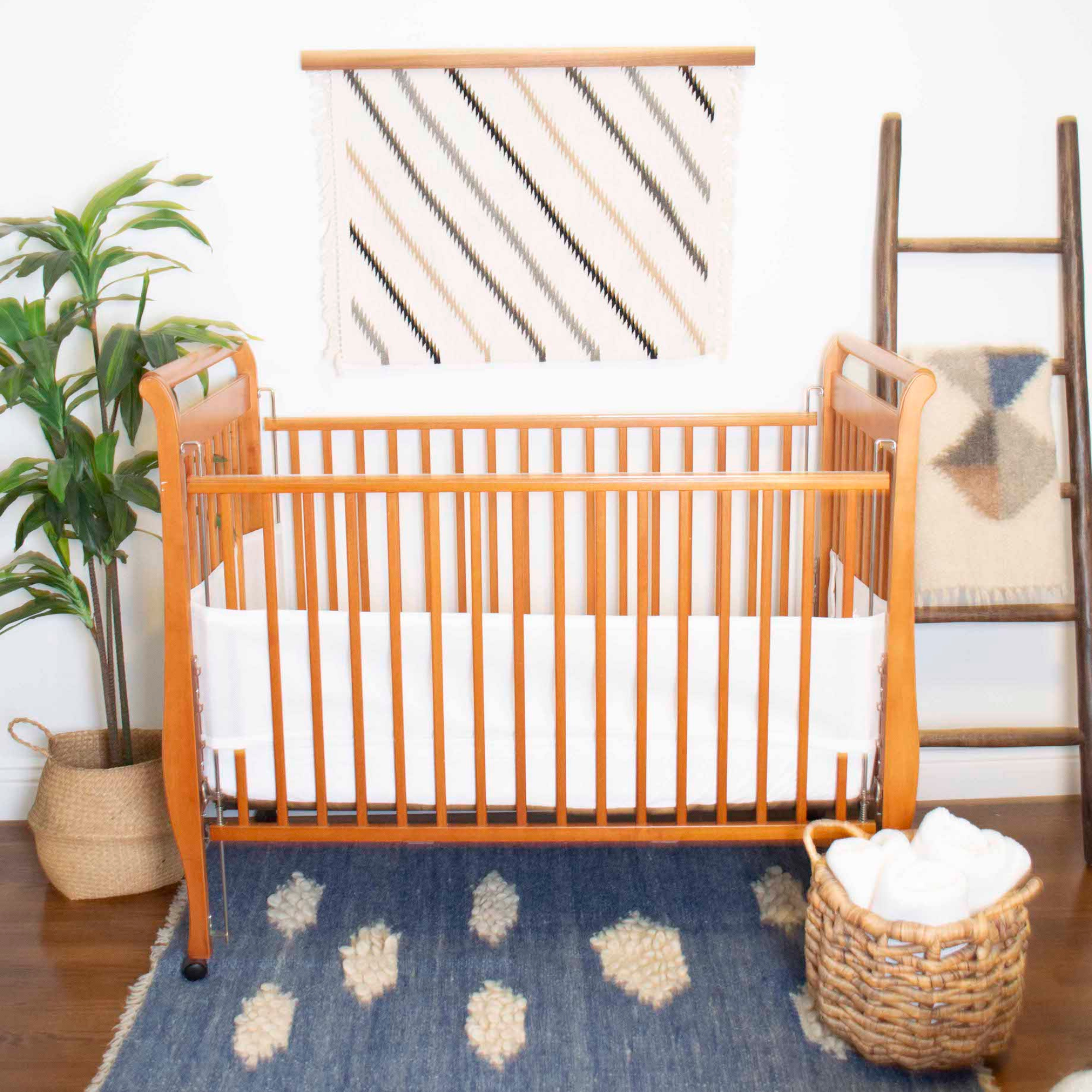 A natural wood crib with white bedding and the Khmara (Cloud) Wool Statement Throw adds warmth, while a plant, woven basket, ladder, and blue rug complete this cozy, inviting nursery space.