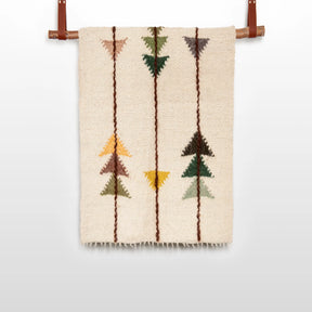 The Lis (Forest) Wool Statement Throw / Baby Blanket features a beige woven design with geometric arrow patterns in brown, green, yellow, and orange, reminiscent of handwoven art, and is displayed hanging from a rod.