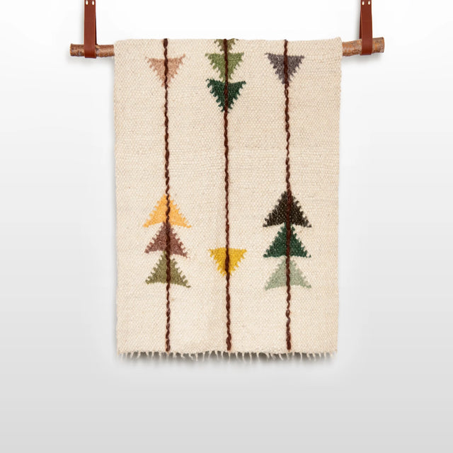 The Lis (Forest) Wool Statement Throw / Baby Blanket features a beige woven design with geometric arrow patterns in brown, green, yellow, and orange, reminiscent of handwoven art, and is displayed hanging from a rod.