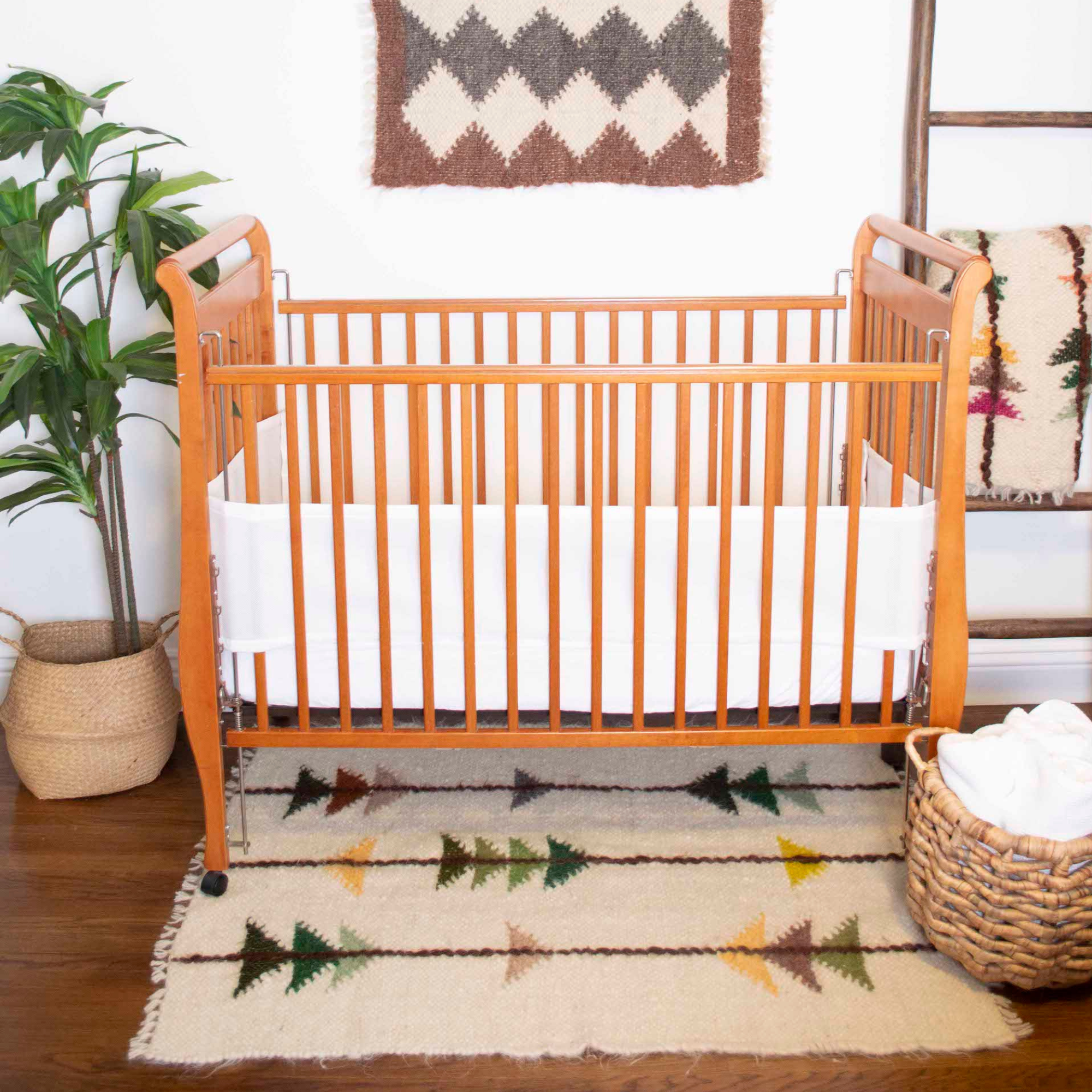 A wooden baby crib with a white bumper sits next to a plant and basket on a tree-patterned rug, topped with the Lis (Forest) Wool Statement Throw / Baby Blanket.