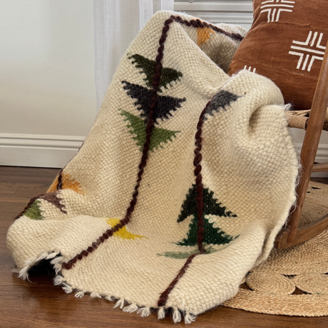The Lis (Forest) Wool Statement Throw / Baby Blanket, crafted from soft Carpathian wool with colorful tree patterns, is draped over a chair beside a brown pillow and woven rug.
