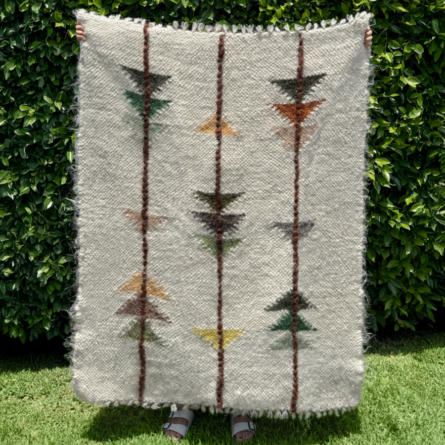 A person holds the Lis (Forest) Wool Statement Throw / Baby Blanket, featuring vertical stripes and geometric patterns, while standing on grass by a leafy hedge.