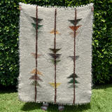 A person holds the Lis (Forest) Wool Statement Throw / Baby Blanket, featuring vertical stripes and geometric patterns, while standing on grass by a leafy hedge.