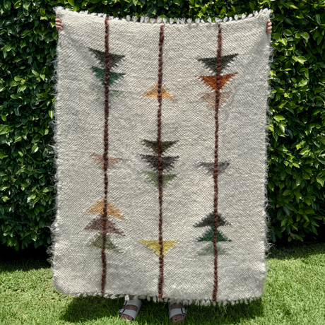A person holds the Lis (Forest) Wool Statement Throw / Baby Blanket, featuring vertical stripes and geometric patterns, while standing on grass by a leafy hedge.