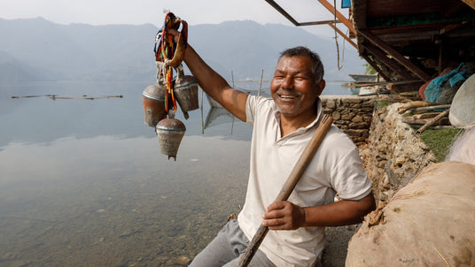 Smiling man by a lake holds up decorated metal bells, with mountains and water in the background.