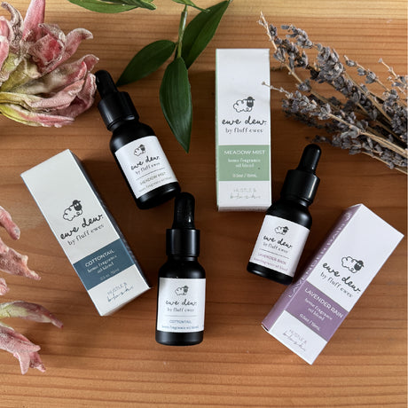 Three bottles of Ewe Dew by Fluff Ewes Meadow Mist Laundry, Home Fragrance + Aromatherapy Essential Oil Blend (0.5 fl oz, Light, Fresh Floral) with matching boxes rest on a wooden surface, surrounded by leaves and dried flowers.