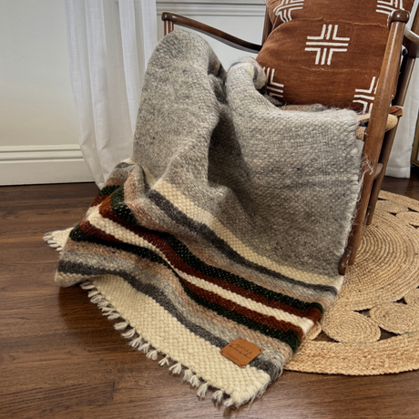 The Mudry (Wise) Wool Statement Baby Blanket in gray stripes is draped over a wooden chair with a patterned pillow, set on a round woven rug.