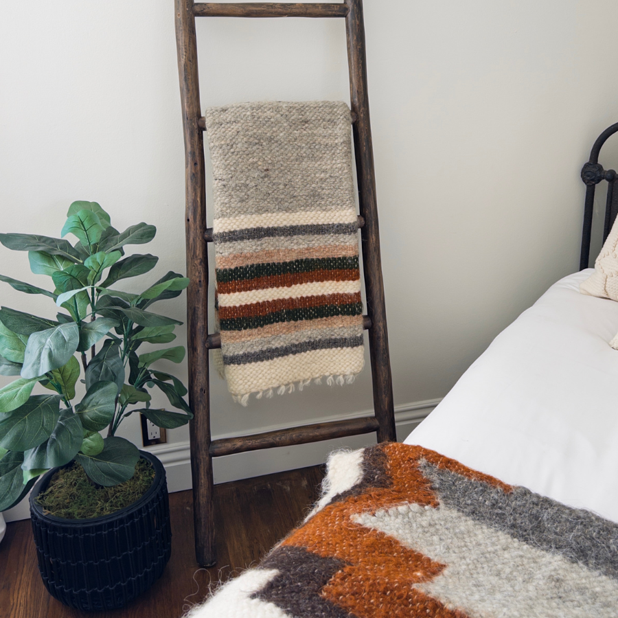 The Mudry (Wise) Wool Statement Baby Blanket hangs on a wooden ladder next to a bed and a potted plant in a cozy room.