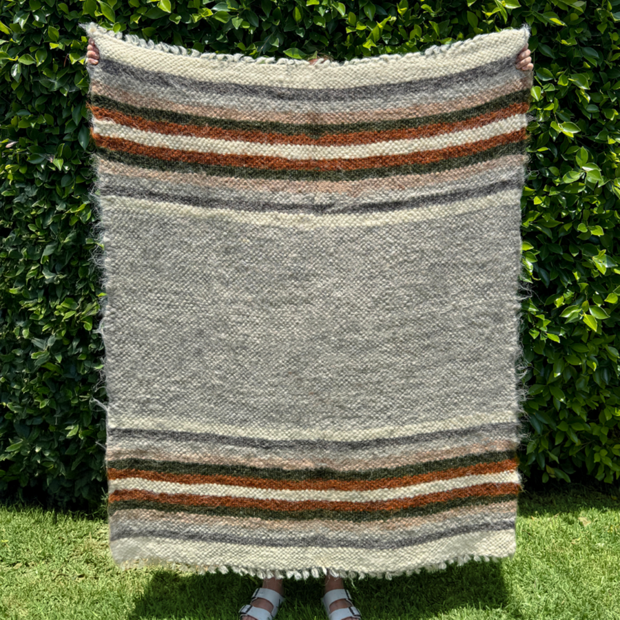 A person holds the Mudry (Wise) Wool Statement Baby Blanket—featuring brown, orange, and dark green stripes—outdoors by a hedge, making it an ideal throw for camping.