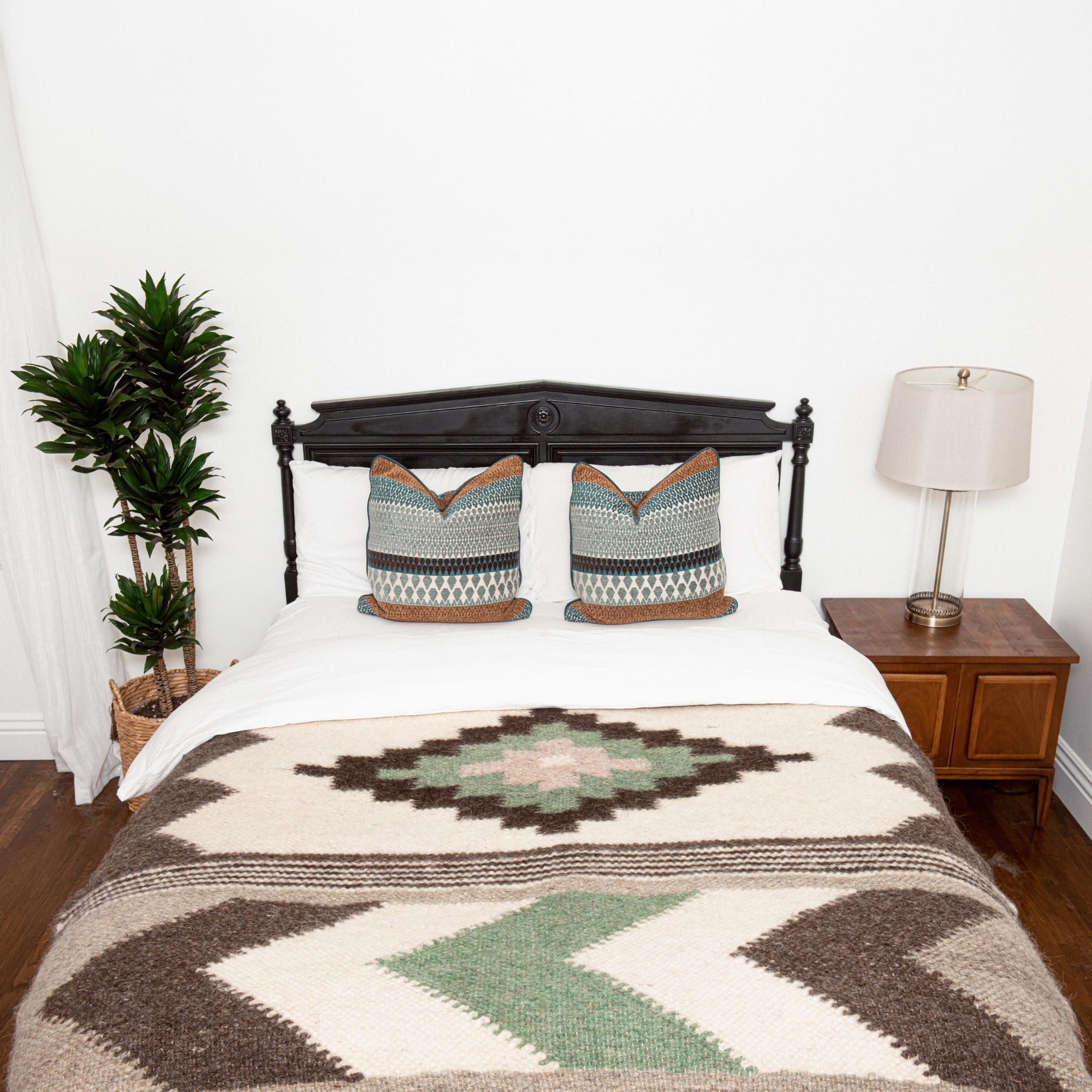 A neatly made bed with patterned pillows and the Myr (Peace) Wool Statement Bed Blanket, flanked by a plant and a lamp on a wooden nightstand.