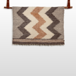 The Myr (Peace) Wool Statement Throw / Baby Blanket, made from all-natural undyed yarn, displays brown, tan, and cream chevron patterns and elegantly hangs on a wooden rod with leather straps.