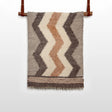 The Myr (Peace) Wool Statement Throw / Baby Blanket, featuring brown, beige, and cream zigzag patterns, hangs on a leather strap rod.