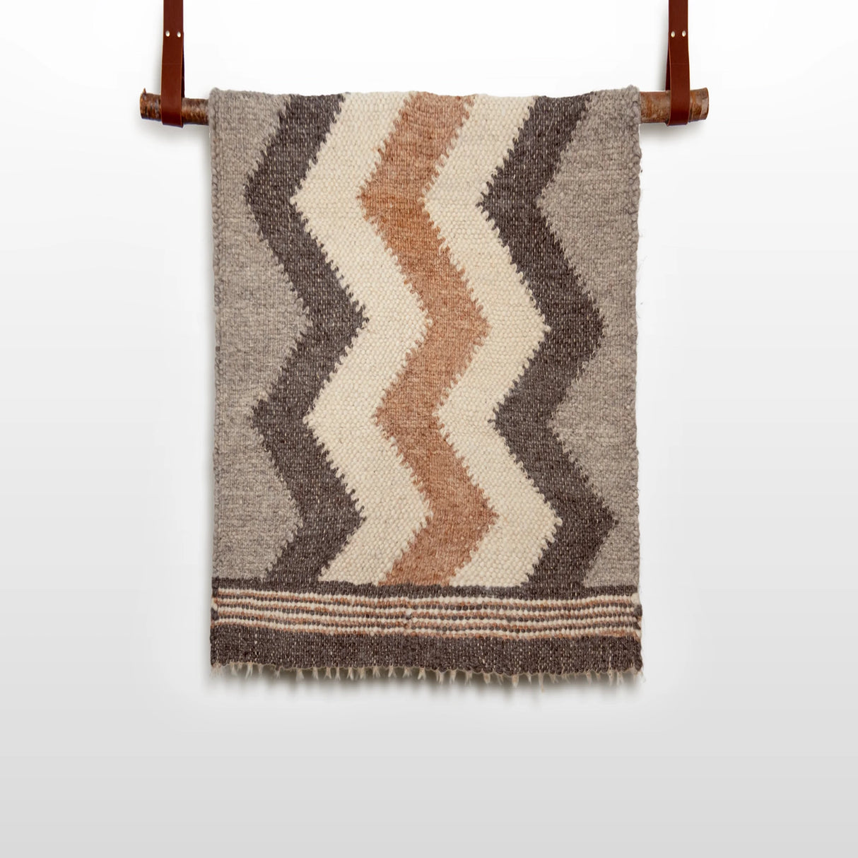 The Myr (Peace) Wool Statement Throw / Baby Blanket, featuring brown, beige, and cream zigzag patterns, hangs on a leather strap rod.