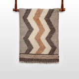The Myr (Peace) Wool Statement Throw / Baby Blanket, featuring brown, beige, and cream zigzag patterns, hangs on a leather strap rod.