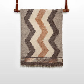 The Myr (Peace) Wool Statement Throw / Baby Blanket, featuring brown, beige, and cream zigzag patterns, hangs on a leather strap rod.