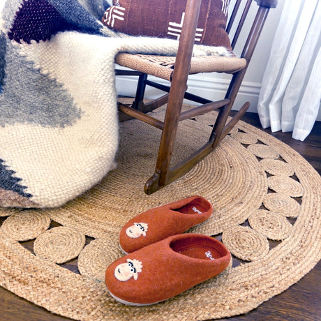 Love Ewe Hand-Felted Organic Wool Slippers for Women (Blush-OG) rest on a woven rug beside a wooden rocking chair draped with a cozy blanket.