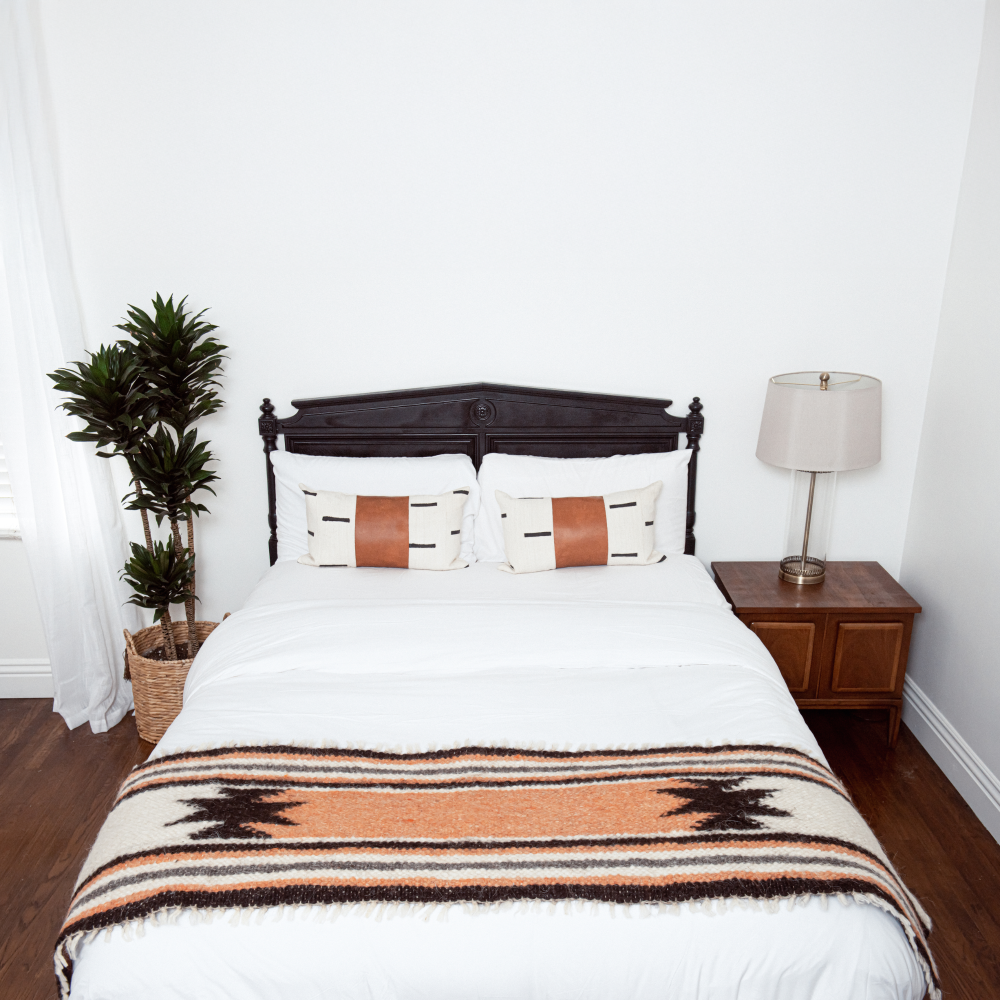 A neatly made bed features patterned pillows and the Opora (Pillar) Wool Bed/Couch Runner, handcrafted from Carpathian sheep wool, beside a lamp on a wooden nightstand and a potted plant.