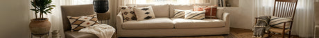 Bright living room with beige sofa, patterned pillows, armchair, wooden rocker, and potted plant by window.