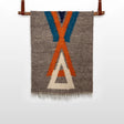 The Podyh (Breathe) Wool Statement Throw / Baby Blanket features a handwoven gray textile with blue, orange, and white geometric patterns, elegantly displayed on a leather rod against a white background.