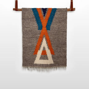 The Podyh (Breathe) Wool Statement Throw / Baby Blanket features a handwoven gray textile with blue, orange, and white geometric patterns, elegantly displayed on a leather rod against a white background.