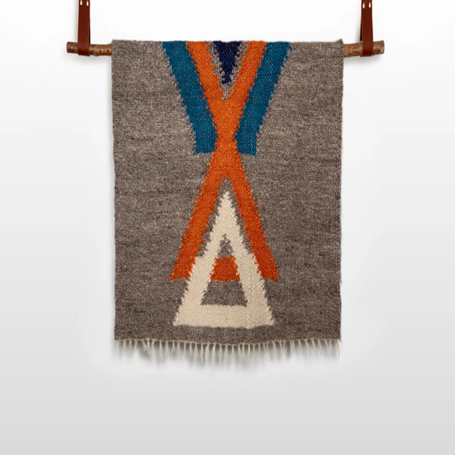 The Podyh (Breathe) Wool Statement Throw / Baby Blanket features a handwoven gray textile with blue, orange, and white geometric patterns, elegantly displayed on a leather rod against a white background.