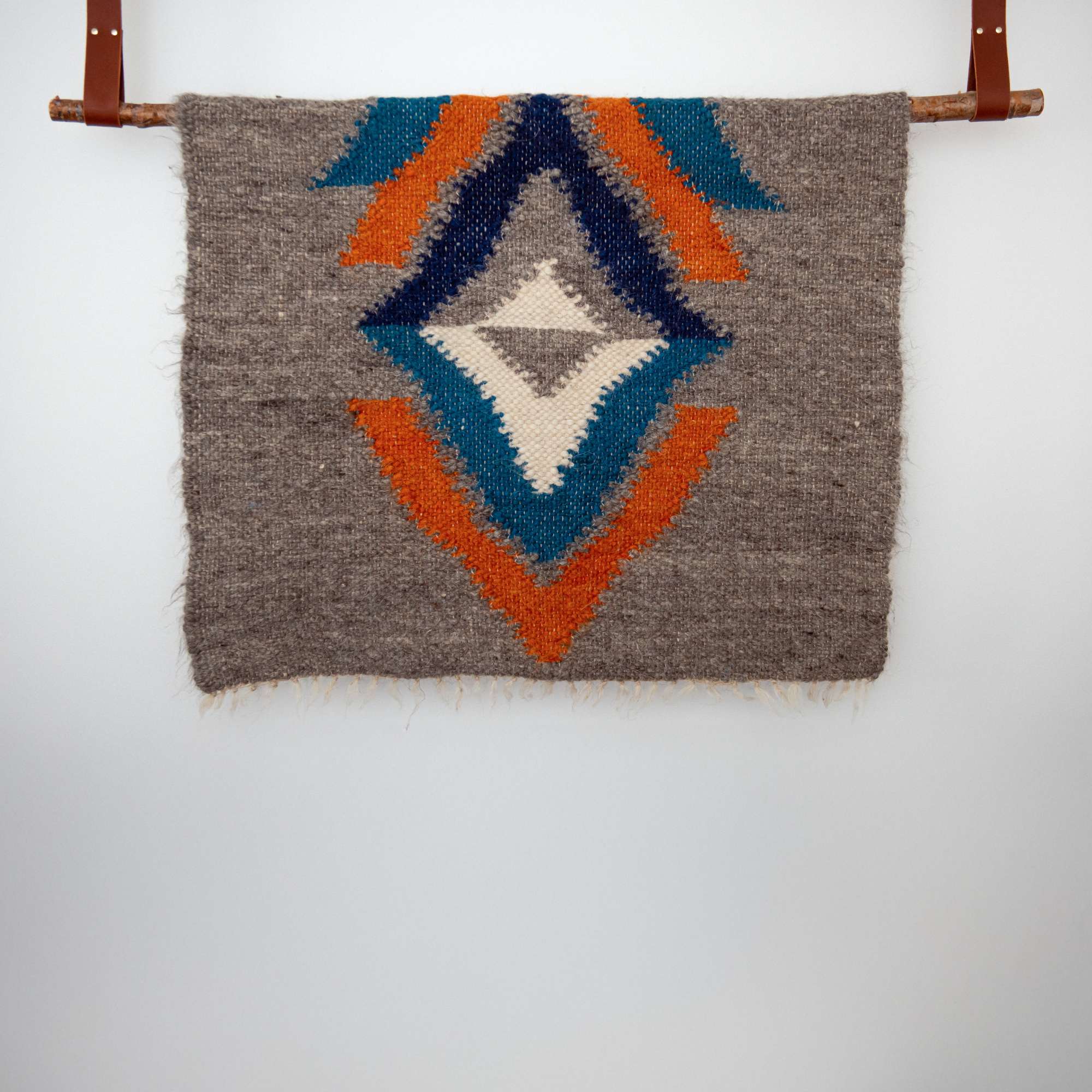 The Podyh (Breathe) Wool Statement Throw / Baby Blanket, featuring geometric patterns in blue, orange, and cream, hangs from a wooden rod on the wall.