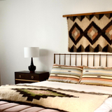 Minimalist bedroom with a metal bed, striped bedding, and a lamp on a nightstand, featuring statement wall decor: the Svitlo (Bright) Wool Wall Hanging.