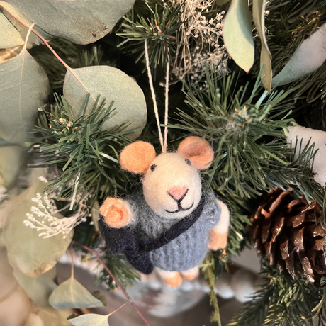The School Boy Mouse is a felt ornament in a blue outfit, handmade in Nepal. It hangs on a Christmas tree with pinecones and eucalyptus, supporting fair trade and adding a charming touch to your décor.