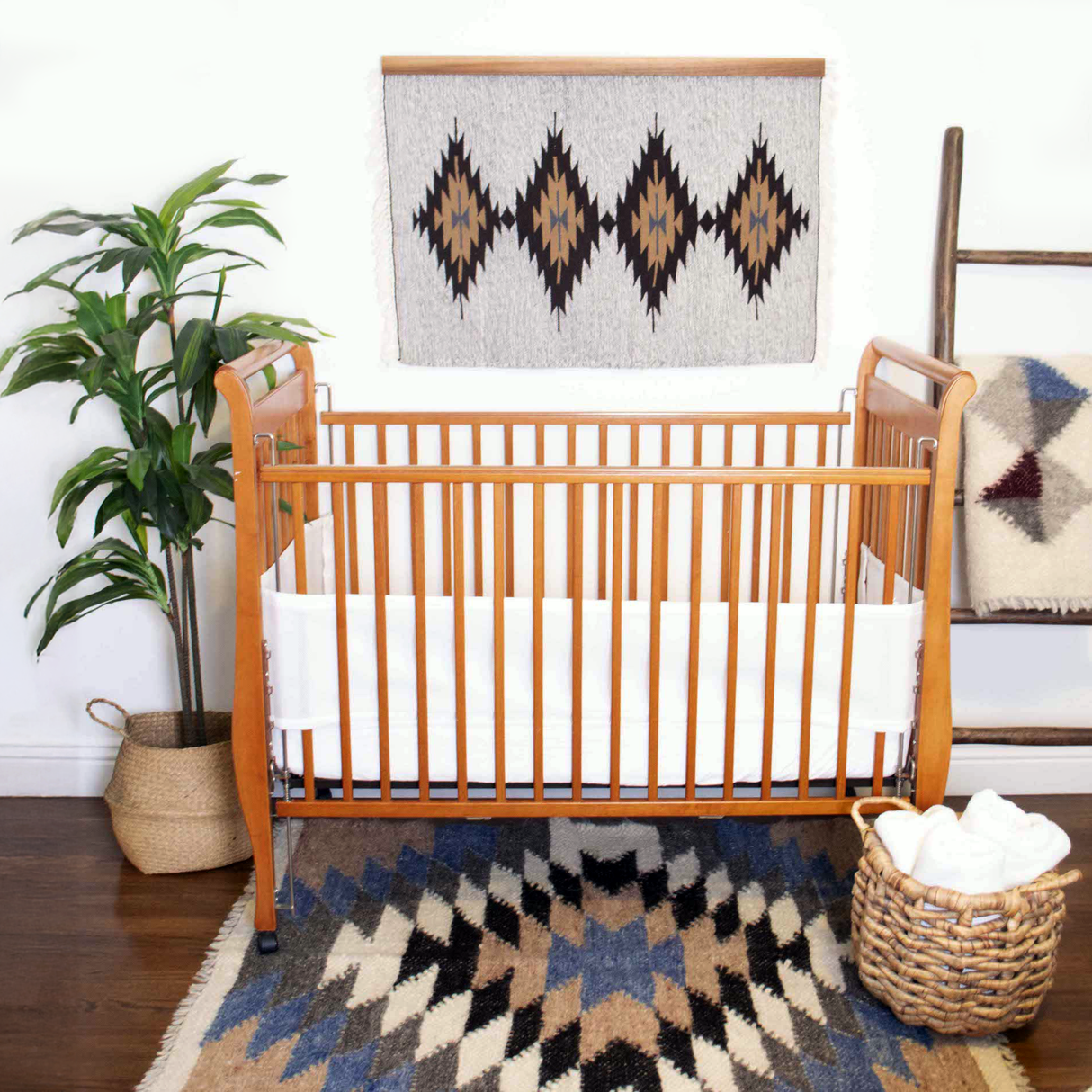Wooden crib in a cozy nursery with a bold Smilyv Wool Rug / Wall Hanging in neutral hues, a potted plant, basket, and geometric wall art.